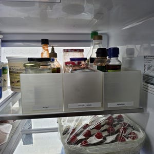 Puede incluir: Interior de un refrigerador con varios alimentos. Frascos, botellas y recipientes están organizados en contenedores blancos etiquetados como "MISC", "JELLY/JAMS" y "ASIAN". Se ve un pastel con cerezas rojas.