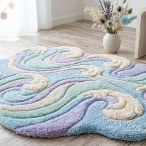 May include: A cloud-shaped rug with a tufted design in pastel colors. The rug features swirling patterns in shades of blue, purple, green, and cream. The rug is placed on a wooden floor, adding a soft and whimsical touch to the room.