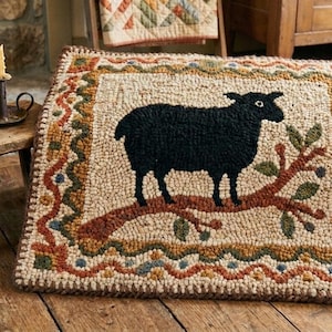 Op de afbeelding: Handgemaakt wollen vloerkleed met een zwart schaap op een bruine tak met groene bladeren. Het vloerkleed heeft een beige achtergrond en een decoratieve rand met oranje, groene en blauwe accenten. Een rustiek woondecoratie-item.