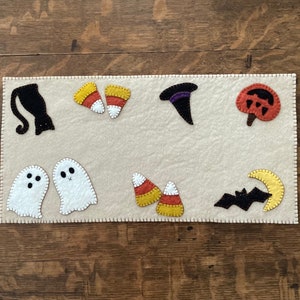 May include: A white felt Halloween-themed doormat with black, orange, yellow, and purple felt appliques. The appliques include a black cat, a witch's hat, a ghost, a bat, and candy corn.