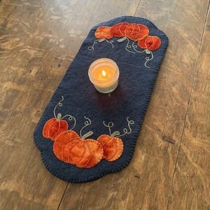 May include: A blue felt table runner with an embroidered design of pumpkins and vines. A lit candle sits in the center of the runner.