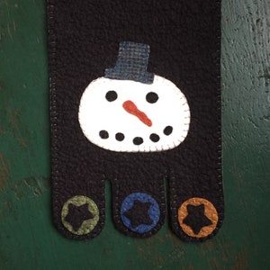 May include: A black felt snowman ornament with a white face, red nose, and black eyes. The snowman has a blue felt hat and three felt feet with stars in green, blue, and orange.