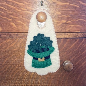 May include: A white felt door hanger with a green felt shamrock and a green felt leprechaun hat. The hat has a gold buckle.