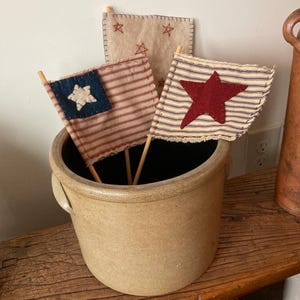 May include: A stoneware crock holds three small, handmade patriotic flags. Two flags feature red and white stripes with a blue star or a red star. The third flag is tan with embroidered stars. The flags are on wooden dowels.