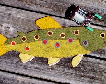 May include: A felt fish wall hanging with a green and yellow body, brown and yellow spots, and a yellow tail fin. The fish is on a wooden background with a vintage fishing reel.