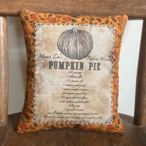May include: A decorative pillow with a pumpkin pie recipe printed on it. The pillow has a cream-colored center with a detailed pumpkin illustration and text. The border is orange with pumpkin and leaf patterns.