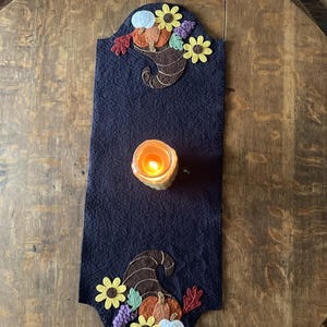 May include: A navy blue felt table runner with autumn-themed appliqués. The design includes a cornucopia, pumpkins, flowers, and a lit candle. The runner is placed on a wooden table, creating a warm, seasonal display.