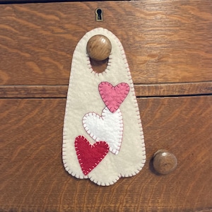 May include: A white felt door hanger with three felt hearts in shades of pink and red. The hearts are stitched together with white thread.