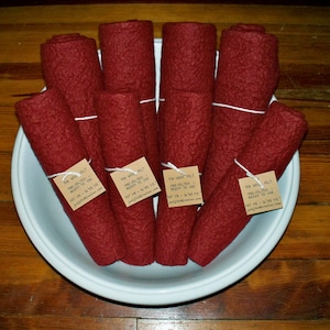 May include: A white bowl filled with eight rolls of red wool felt. Each roll is tied with twine and has a brown label that reads "100% Wool Felt Pre-Felted - Ready to Use Fat 1/8 - 18 x 18" with a website address.