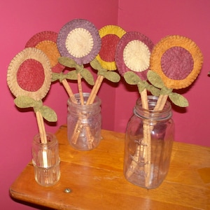May include: A bouquet of felt flower crafts with cinnamon stick stems in glass jars. The flowers are in shades of red, orange, brown, and purple.