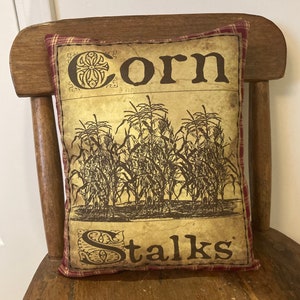 May include: A decorative pillow with a red and white plaid border. The pillow features a vintage-style illustration of corn stalks with the text "Corn Stalks" in a decorative font.