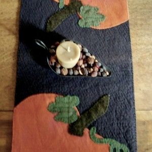 May include: A dark blue felt table runner with two orange felt pumpkins and a metal candle holder filled with brown and white decorative stones. The candle holder is in the center of the runner and the pumpkins are on either side.