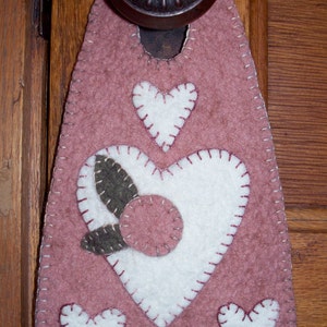May include: A pink felt doorstop with a white heart design and a smaller green leaf design. The doorstop is shaped like a heart and has a scalloped edge.