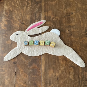 May include: A white felt bunny rabbit with pink ears and a white tail. The bunny is laying on a wooden surface with the word "SPRING" spelled out in colorful wooden blocks.