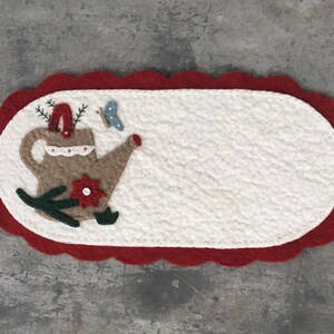 May include: A white felt oval-shaped doormat with a red scalloped border. The doormat features an embroidered design of a brown watering can with a red flower and green leaves.