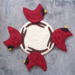 May include: Four felt cardinal birds are arranged around a cream-colored felt circle with brown felt branches. The birds are red with black eyes and yellow beaks.