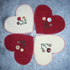 May include: Four heart-shaped felt coasters arranged in a cloverleaf pattern. Two are cream-colored, two are red. Each has a green embroidered sprig and decorative buttons. The edges are stitched with red thread.