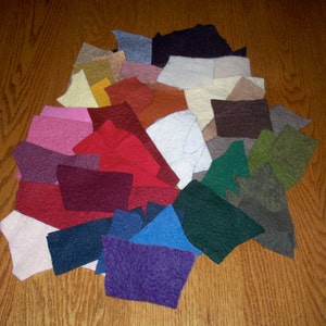 May include: Assortment of felt fabric swatches in various colors, including red, green, blue, purple, brown, white, and pink. The swatches are arranged in a fan shape on a wooden surface.