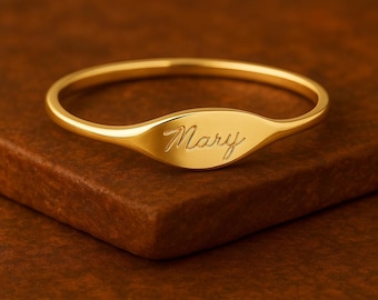May include: A gold-colored ring with the name "Mary" engraved in a cursive font. The ring has a smooth, polished finish and a rounded, oval-shaped face. The ring is set on a brown surface.
