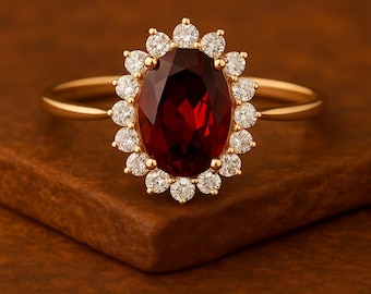 Puede incluir: Anillo de oro con una gran gema roja oscura de corte ovalado, rodeada por un halo de pequeñas gemas blancas redondas. El anillo tiene una fina banda de oro y una montura dorada.