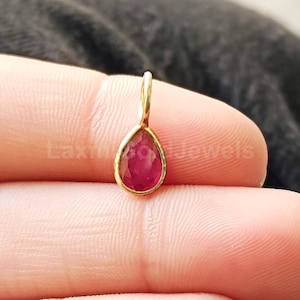 May include: A teardrop-shaped pendant featuring a faceted red ruby gemstone set in a gold bezel frame with a small attached bail at the top. The pendant is positioned against a dark textured background with the text LaxmiGoldJewels visible.