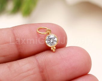 May include: A small, gold-colored charm with a clear, round gemstone. The charm has a small loop at the top and three small gold beads at the bottom. The charm is held in a person's hand.