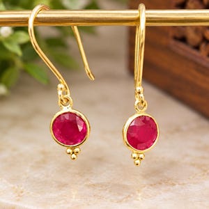 Peut inclure: Une paire de boucles d'oreilles pendantes en or avec des pierres rondes à facettes rose foncé serties dans des montures en or. Chaque boucle d'oreilles est suspendue à un crochet doré et comporte trois petites sphères dorées à la base.