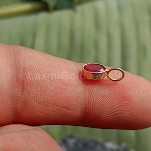 Puede incluir: Un pequeño colgante de oro con una vibrante piedra preciosa roja. La piedra de forma ovalada está engastada en un bisel de oro, y el colgante tiene un pequeño lazo para una cadena. La joya se muestra en un dedo.