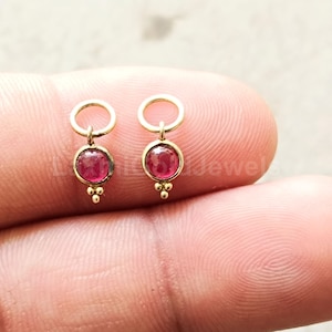 Peut inclure: Une paire de boucles d'oreilles en or composée d'une boucle supérieure circulaire reliée à une pierre précieuse rose foncé sertie clos. Sous chaque pierre, trois petites sphères dorées forment un motif triangulaire délicat et symétrique.