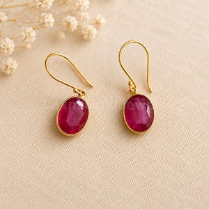Puede incluir: Un par de pendientes colgantes de oro con piedras preciosas ovaladas y facetadas de color rosa intenso. Cada pendiente cuelga de un sencillo gancho de oro, mostrando un engaste de metal pulido que enmarca las piedras rojo-rosadas.