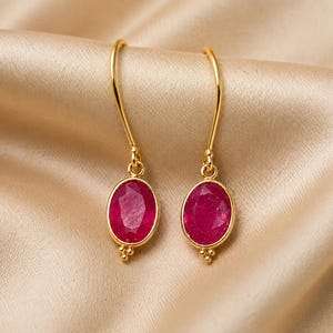 Peut inclure: Une paire de boucles d'oreilles pendantes en or avec des pierres précieuses rubis rose foncé à facettes de forme ovale. Chaque boucle est suspendue à un crochet doré incurvé et présente un petit groupe décoratif de trois minuscules perles dorées.