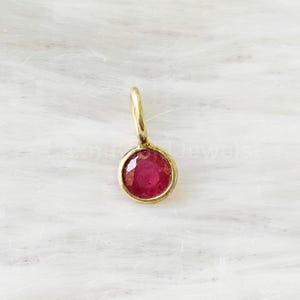 Könnte beinhalten: Ein kleiner, runder Anhänger mit einem facettierten, tief rosa-roten Edelstein, der in einer glatten, polierten goldfarbenen Fassung sitzt. Der Anhänger verfügt über einen kleinen, passenden goldfarbenen Ring an der Oberseite.