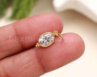 Puede incluir: Un colgante ovalado dorado con una piedra preciosa clara y facetada. El colgante tiene un pequeño lazo en la parte superior para sujetarlo a una cadena o pulsera. La piedra está engastada en un bisel dorado.
