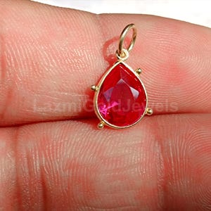May include: A teardrop-shaped pendant with a vibrant red gemstone set in a gold-colored frame. The pendant has a small loop at the top for a chain and small decorative accents. The gemstone is faceted, catching the light.