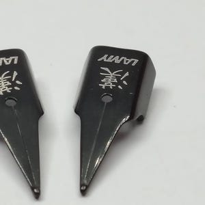 Puede incluir: Primer plano de dos plumines negros para pluma estilogr&aacute;fica Lamy. Los plumines tienen la marca "LAMY" y un car&aacute;cter asi&aacute;tico grabado. Los plumines est&aacute;n sobre un fondo blanco.