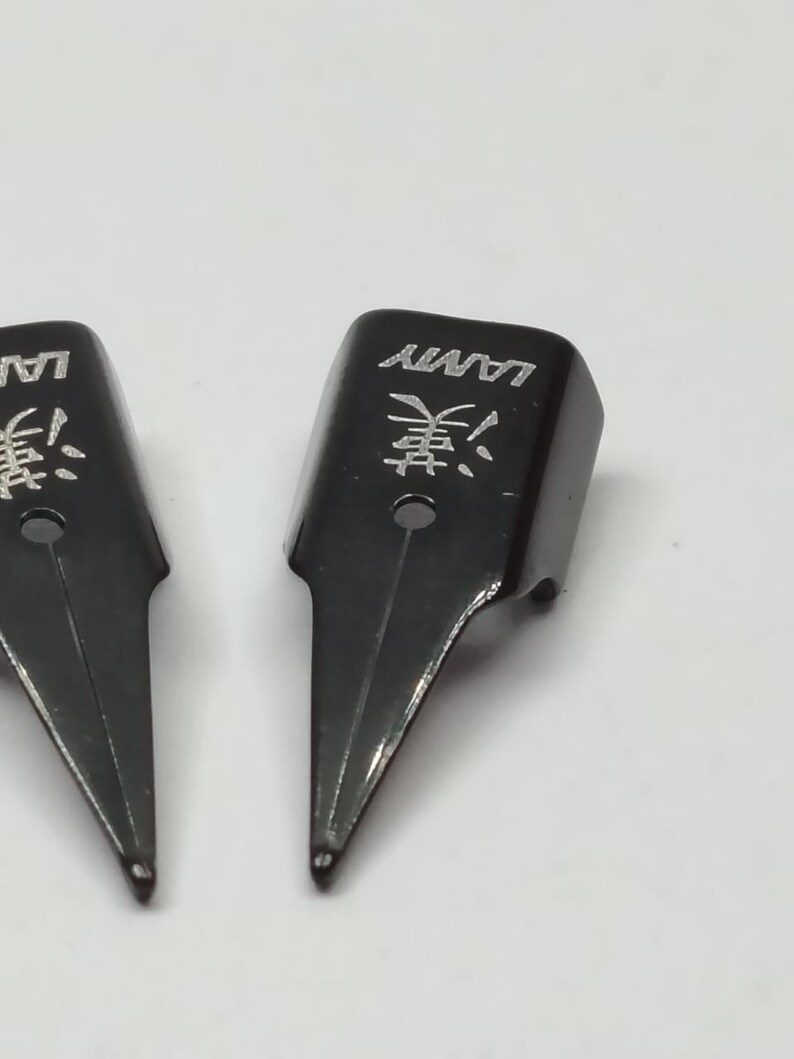 Puede incluir: Primer plano de dos plumines negros para pluma estilogr&aacute;fica Lamy. Los plumines tienen la marca "LAMY" y un car&aacute;cter asi&aacute;tico grabado. Los plumines est&aacute;n sobre un fondo blanco.