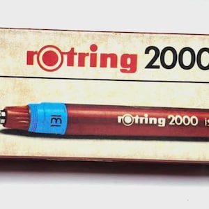 Puede incluir: Un estilógrafo técnico Rotring 2000 isograph con un cuerpo rojo y una banda azul marcada con "131". El bolígrafo está junto a su tapa y una caja de cartón con el logotipo de Rotring y "2000 isograph" impreso.