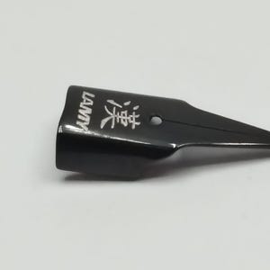 Puede incluir: Una plumilla negra con la palabra "LAMY" impresa en blanco en el lateral. La plumilla tambi&eacute;n tiene caracteres chinos y un peque&ntilde;o agujero. La plumilla est&aacute; sobre un fondo blanco.