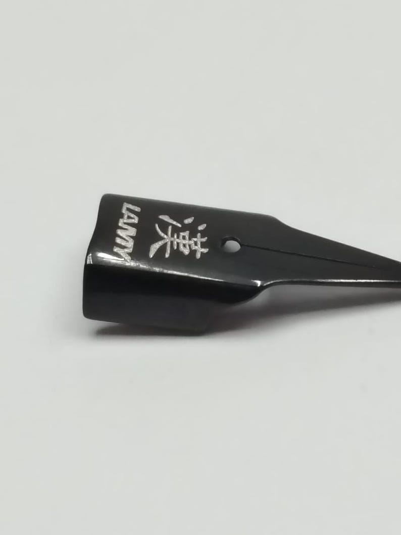 Puede incluir: Una plumilla negra con la palabra "LAMY" impresa en blanco en el lateral. La plumilla tambi&eacute;n tiene caracteres chinos y un peque&ntilde;o agujero. La plumilla est&aacute; sobre un fondo blanco.