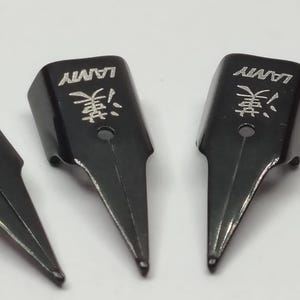Puede incluir: Primer plano de plumines negros. Cada plum&iacute;n tiene la palabra "LAMY" y un car&aacute;cter japon&eacute;s grabado. Los plumines son de metal y tienen una punta. El fondo es de color claro.