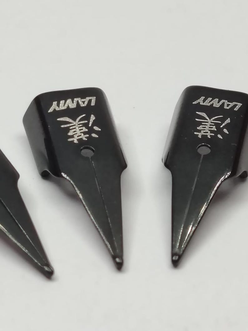 Puede incluir: Primer plano de plumines negros. Cada plum&iacute;n tiene la palabra "LAMY" y un car&aacute;cter japon&eacute;s grabado. Los plumines son de metal y tienen una punta. El fondo es de color claro.