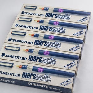 May include: A stack of Staedtler Marsmatic technical pens in their original packaging. The pens are dark blue with silver accents and feature the text "marsmatic" and "DURANITE pro-fum". The boxes are white and blue.