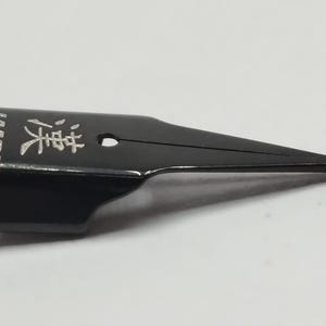 Puede incluir: Una plumilla de pluma estilogr&aacute;fica negra con la palabra "LAMY" y caracteres japoneses grabados en plata. La plumilla tiene un dise&ntilde;o elegante y moderno con una punta fina.