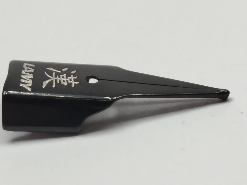 Puede incluir: Una plumilla de pluma estilogr&aacute;fica negra con la palabra "LAMY" y caracteres japoneses grabados en plata. La plumilla tiene un dise&ntilde;o elegante y moderno con una punta fina.
