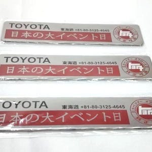 May include: Three rectangular Toyota stickers. Each sticker has the Toyota logo, Japanese text, and a phone number. The stickers are silver with red and white text and a circular logo on the right.