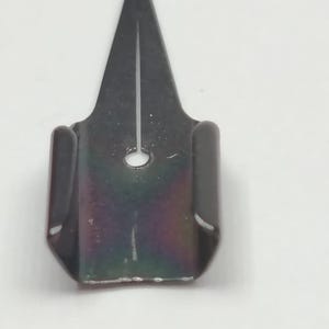 Puede incluir: Un portaplumas de metal gris oscuro para una pluma de caligraf&iacute;a. El soporte tiene una punta afilada y un peque&ntilde;o agujero cerca de la base. El metal tiene un ligero brillo iridiscente, que refleja colores como el p&uacute;rpura y el verde.