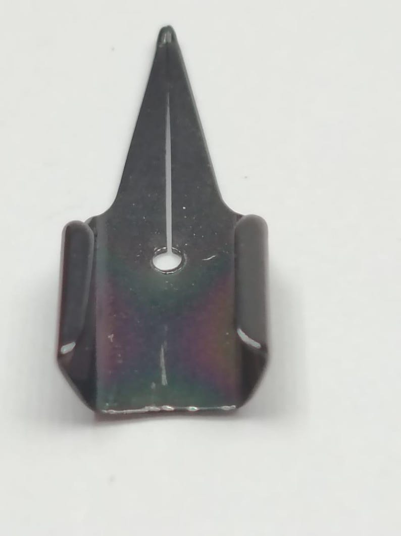 Puede incluir: Un portaplumas de metal gris oscuro para una pluma de caligraf&iacute;a. El soporte tiene una punta afilada y un peque&ntilde;o agujero cerca de la base. El metal tiene un ligero brillo iridiscente, que refleja colores como el p&uacute;rpura y el verde.