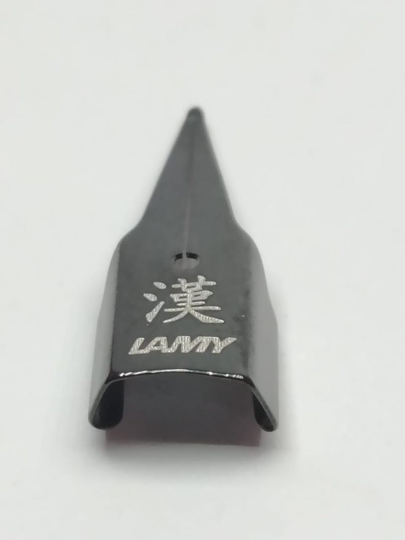 Puede incluir: Una plumilla negra con una punta afilada. La plumilla tiene la palabra "LAMY" y un car&aacute;cter chino grabado. La plumilla es de metal y tiene un acabado brillante.