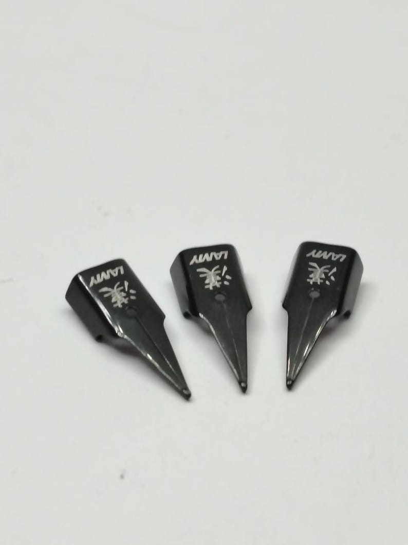 Puede incluir: Tres plumines negros de pluma estilogr&aacute;fica Lamy est&aacute;n colocados sobre una superficie blanca. Cada plum&iacute;n tiene el logotipo y el nombre de la marca Lamy. Los plumines est&aacute;n en &aacute;ngulo hacia la parte inferior derecha.