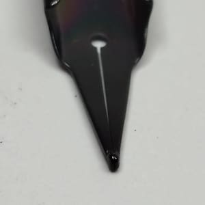 Puede incluir: Primer plano de una plumilla de pluma estilogr&aacute;fica negra. La plumilla tiene una punta fina con una ranura en el centro. La plumilla es de metal y tiene un acabado brillante. El fondo es una superficie blanca lisa.